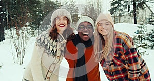 Winter vacation together. Three multiethnic happy friends wave, smile at camera at beautiful snowy forest slow motion.