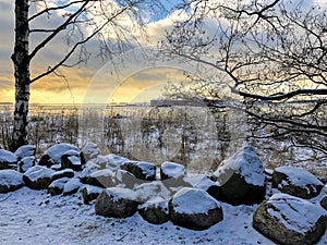 Winter view over the Baltic Sea with snow covered stones on the shore and trees under the sky with sun lighted clouds