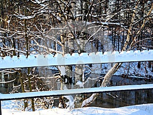 Winter view of shaped by hand snow cover of the bridge railing with the birch tree and still river behind