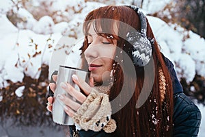 Winter. Woman with red hair wearing ear muffs. Girl drinking hot tea or coffee iron insulated cup