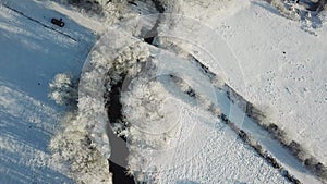 Winter woodland in frost top aerial view. Trees on riverbank. Small creek in forest. Rural dirt road. Snow covered field landscape