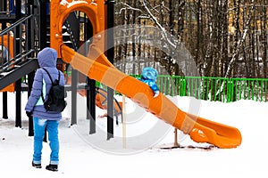 Wintertime. Mom watching little kid sliding the slide on the playground