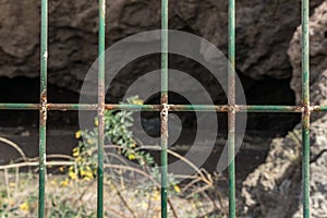 Wire mesh fence closeup photo. Green bush and huge cave in blurred background. Thin pillars. Decorative board front of garden.