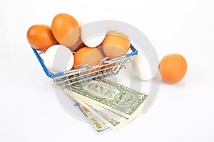 A wire shopping basket filled with white and brown eggs, placed beside rolled dollar bills and an egg, on a reflective white