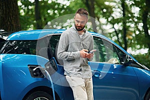 Wireless device, using smartphone. Handsome man is standing outdoors near electric automobile and waiting for charging