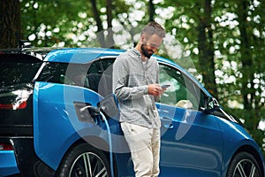 Wireless device, using smartphone. Handsome man is standing outdoors near electric automobile and waiting for charging