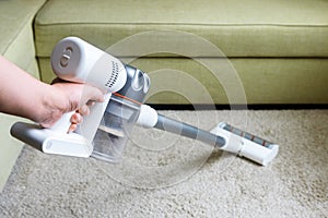 Wireless vacuum cleaner used on carpet in room. Housework with new white hoover. Person holds modern vacuum cleaner by sofa