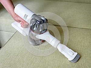 Wireless vacuum cleaner used on sofa in room. Housework with new upright hoover and nozzle. Person holds modern white vacuum