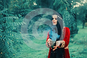 Wise fortune-teller in silk red scarlet dress and gold decoration stands alone in forest with magic bowl, calling