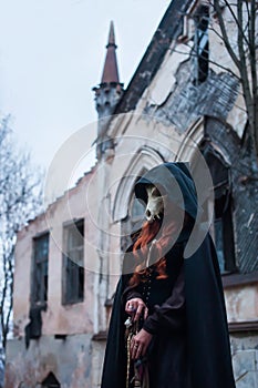 A witch in a black cloak with a hood with red hair stands at dusk against the background of an old abandoned castle