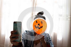 Witch holding a pumpkin balloon with her hand, covering her face and taking a photo with mobile phone. Young woman celebrating