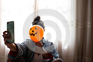 Witch holding a pumpkin balloon with her hand, covering her face and taking a photo with mobile phone. Young woman celebrating