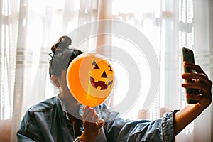 Witch holding a pumpkin balloon with her hand, covering her face and taking a photo with mobile phone. Young woman celebrating