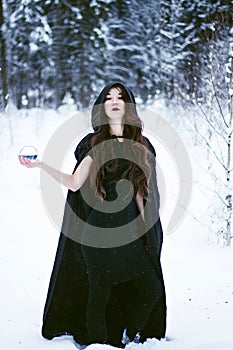 Witch or woman in black cloak with glass ball in white snow forest