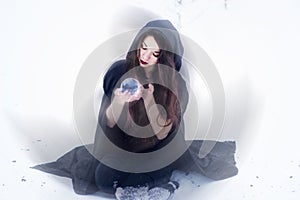 Witch or woman doing magic in black cloak with glass ball in white snow forest