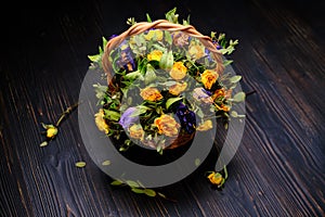 Withered flowers with fallen leaves on a wooden black background. Wicker basket with dry yellow and blue roses. Autumn flower