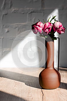 Withered pink roses in a vase in the hard rays of the sun on a table against a background of white brick