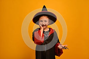 Wizard girl holding toy spider and looking at camera on yellow background. Surprised wizard kid holding hand on cheek.