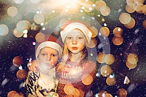 Wo siblings celebrating christmas eve wearing a Santa hat and holding sparklers in the hands. Decorated tree and bokeh in the back