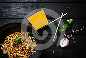 Wok. Udon stir fry noodles with chicken and vegetables in wok pan on black wooden background. With a box for noodles