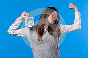 Woken up by alarm clock sleepy student girl holding it in hand. Blue background. Early 7 o'clock in morning