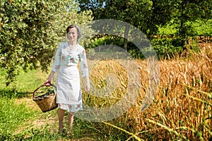Woman in 40s clothes walks in the Italian countryside, next to a wheat field, carrying a basket of cherries