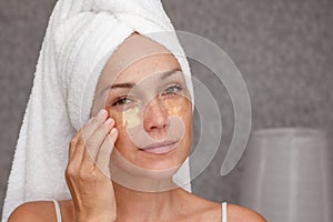 Woman of 40s after shower with towel on head with gold patches under eyes on face