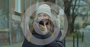 Woman activates smartphone digital virtual app voice assistant recording audio messages at outdoor. Young girl in hat