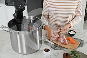 Woman adding rosemary to meat near pot with sous vide cooker at table, closeup. Thermal immersion circulator