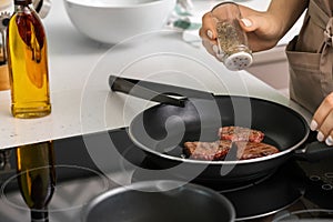 Woman adding spices to meat on frying pan in kitchen, closeup