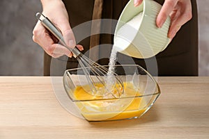 Woman adding sugar to whisked eggs at wooden table, closeup photo