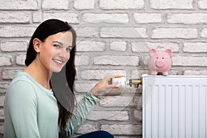 Woman Adjusting Thermostat With Piggy Bank On Radiator