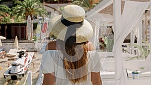 Woman, adorned in a light dress and straw hat, walks along a wooden pier by the sea, embracing the joys of vacation