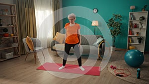 A woman of age takes care of her health, does exercises at home. A grayhaired woman in sportswear and glasses leans