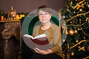 A woman aged dressed in a green sweater reads a book in glasses sitting in a rocking chair near a Christmas tree