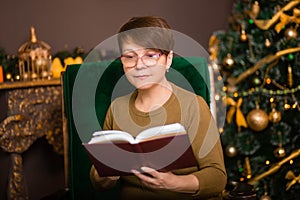 A woman aged dressed in a green sweater reads a book in glasses sitting in a rocking chair near a Christmas tree