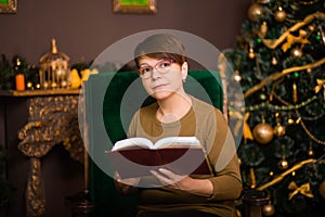 A woman aged dressed in a green sweater reads a book in glasses sitting in a rocking chair near a Christmas tree