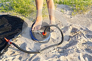 A woman with air foot pump pumps an inflatable mattress or air bed at sandy beach. Foot inflates air mattress with foot