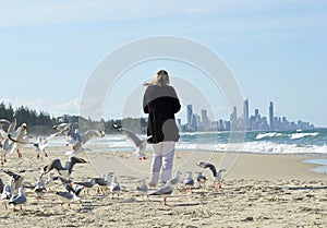 Woman alone walking on beach surrounded by flock sea birds & distant city life
