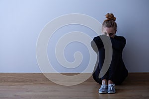 Woman with anxiety disorder wearing dark clothes sitting on the floor, copy space on empty wall