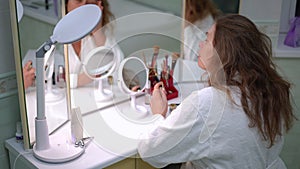 A woman applies cream to her face with her finger while sitting in a white bathrobe in front of a three-leaf makeup