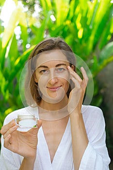 A woman applies cream under her eyes with massage movements and holding a jar.
