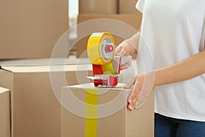 Woman applying adhesive tape on box with dispenser indoors, closeup