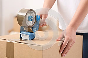 Woman applying adhesive tape on box with dispenser indoors, closeup