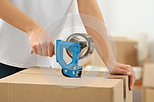 Woman applying adhesive tape on box with dispenser indoors, closeup