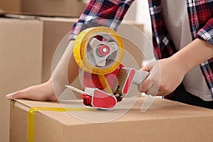 Woman applying adhesive tape on box with dispenser indoors, closeup