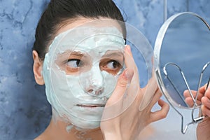 Woman is applying alginate mask in bathr with foam looking at small mirror.