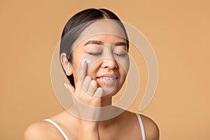 woman applying a anti age or moisturizer face cream on her face, smiling sweetly standing on beige background