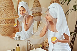 Woman Applying Clay Mask. Wrapped In Bath Towel Model Looking At Mirror And Using Skin Care Product For Derma. photo