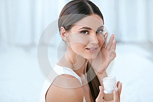 Woman Applying Facial Cream On Face Skin In White Interior